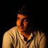 Warm lighting portrait of a thoughtful man with dark hair, wearing a beige shirt, illuminated softly against a black background, emphasizing contemplative mood and detail.
